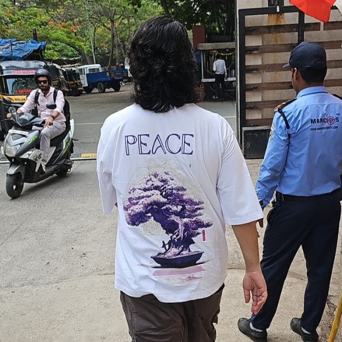 Person wearing a white shirt with 'PEACE' printed on the back, walking on a sidewalk with a street scene in the background.